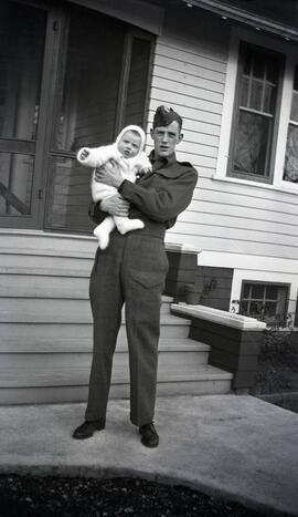 Gordon Headridge holding baby in front of likely Essondale Cottage 118